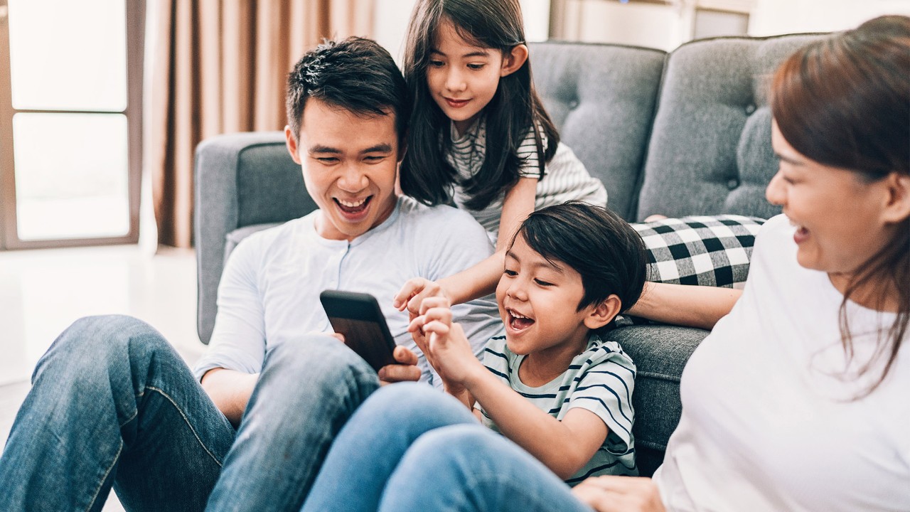 kids with parents on sofa, smiling and looking at smart phone; image used for Manage Your Portfolio for HSBC Singapore Wealth