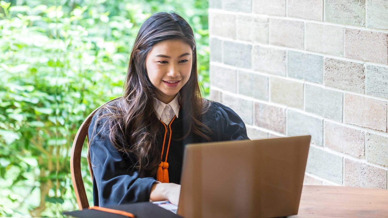 Lady in graduation gown using laptop.