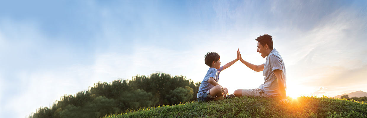 A father and son giving each other a high five on meadow; image used for HSBC Singapore Emerald Life Legacy Plan.